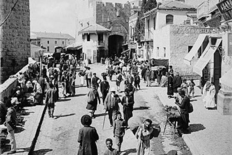 Crowded early twentieth-century street in Jerusalem's Old City near Jaffa Gate, with pedestrians, shops, and the Fr. Vester and Company storefront visible.